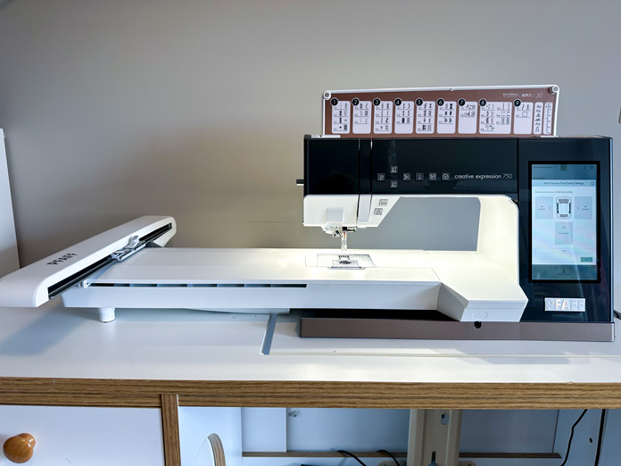 A picture of a deep charcoal and white sewing /embroidery machine with a large computer screen, turned on, waiting to be used. A white embroidery arm attached.