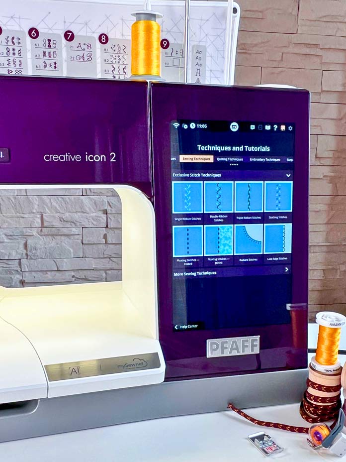 Close-up of the PFAFF Creative Icon 2 sewing and embroidery machine 10.1-inch capacitive touchscreen displaying built-in Exclusive Stitch Techniques Tutorials, including Single, Double, and Triple Ribbon Stitches. A spool of yellow Mettler thread and a wooden spool with decorative 8 mm brown decorative ribbon, and a PFAFF 6D Dynamic Spring Foot and 2A Fancy Stitch Foot are visible nearby.