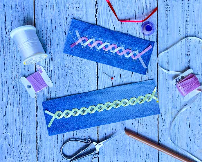 Two stitched denim samples showcasing the Double Ribbon Stitches created using the PFAFF creative icon 2. The left sample features ribbon stitch #5 a yellow ribbon with built-in decorative accents forming crisscrossed layers, the right sample features ribbon stitch #3 pink and white sheer ribbons. Various sewing tools, pink ribbons, and bobbins are arranged around the denim pieces on a light wood background, emphasizing the creative process and embellishment details.