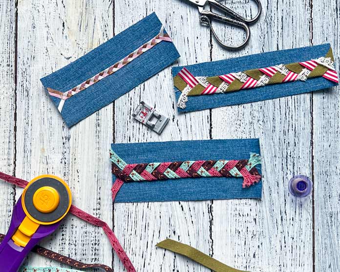 A flatlay of three denim fabric samples featuring intricate Exclusive Triple Ribbon Stitches #4, #8 and #10 created with the PFAFF creative icon 2. Each sample shows a unique color combination of ribbons stitched in decorative patterns using the Triple Ribbon Stitch technique. Surrounding the samples are sewing tools, including a rotary cutter, a spool of green ribbon, a presser foot, and a purple bobbin, all arranged on a textured white wooden surface.