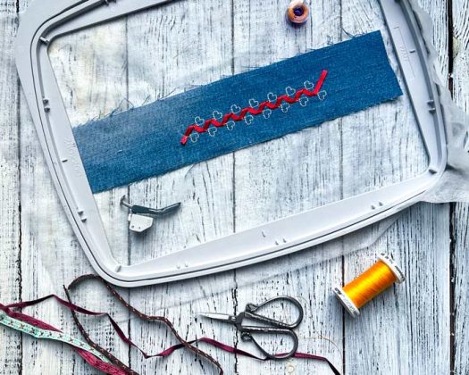 A stitched sample of Single Ribbon Stitch 8.1.6 using 5 mm red ribbon on denim, hooped in the Elite Hoop 260 x 200 mm with the PFAFF creative icon 2 in Embroidery Mode. The sample lies flat on whitewashed wooden surface alongside a yellow spool of Mettler 40wt thread, a bobbin, scissors, a 6D Dynamic Spring Foot, and two types of decorative ribbons, suggesting a creative and practical workspace setup.