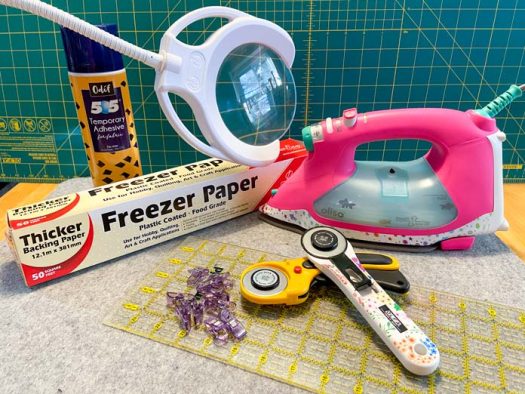 A package of freezer paper, two rotary cutters, some clips, a ruler, an iron, and can of adhesive spray sit on top of a grey wool pressing mat with a green cutting mat in the background.