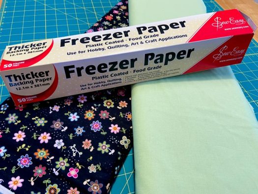 A box of freezer paper sits on top of two bolts of fabric. One fabric is black with small flowers, and the other is light green. Everything sits on top of a green cutting mat.