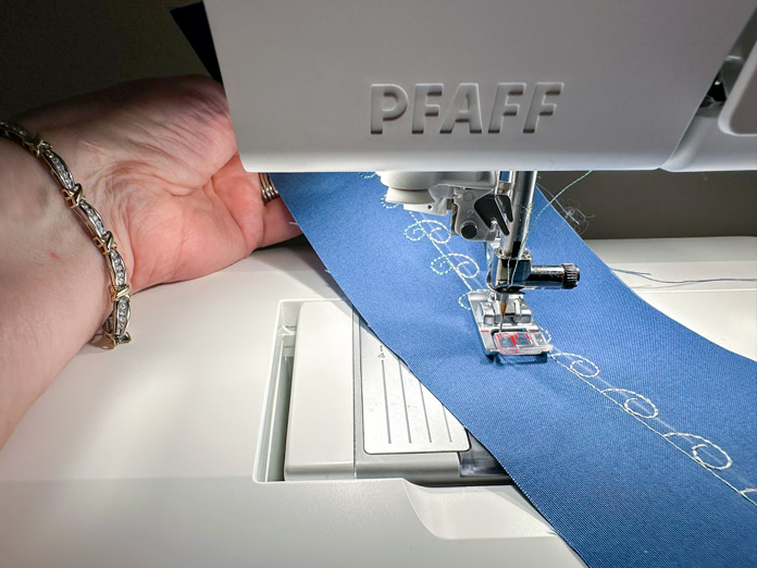 A blue piece of fabric with a decorative swirl stitch (1 of 2), in a light green, a straight stitch in the middle, with the swirl going to the right. Followed by a second swirl stitch in a different green thread, following the straight line in the center with the swirl, going to the left. Finished look, a straight line and the swirls in 2 colors going out to the left and right.