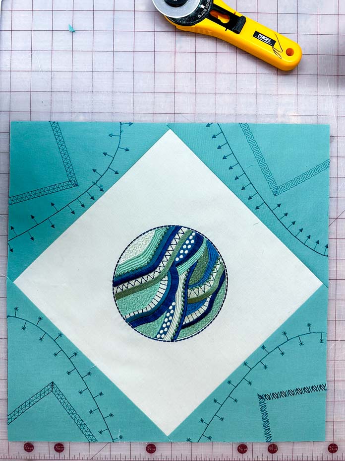 Shades of green fabric pieced together. The center square a light green, has an embroidered filled circle of mutiple green shades of embroidery thread. 