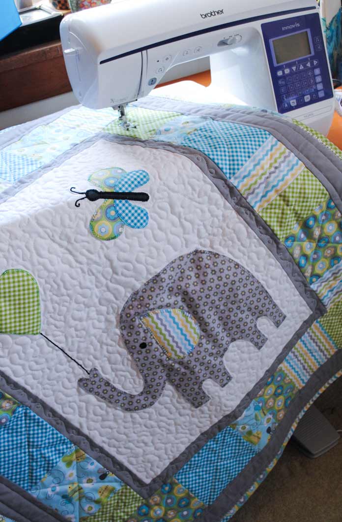 A white, grey, green, and blue baby quilt featuring an elephant motif is shown displayed on the bed of a white and blue NQ900 sewing machine from Brother. Brother NQ900 sewing machine