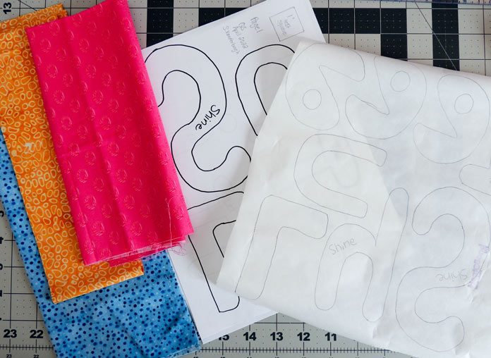 Pieces of pink, orange and blue fabric next to applique letters printed on HeatnBond Lite; Inspira Fast & Easy Tear-A-Way Light