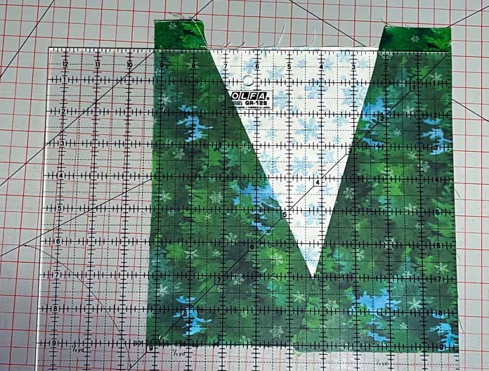 A square quilting ruler lays on top of an untrimmed tree quilt block comprised of an inverted triangle (point down) in white printed fabric and green fabric on both sides as background; the block is on a gray cutting mat with red grid lines