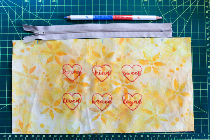 A yellow rectangular piece of fabric with 6 hearts embroidered with words in red thread lays below a gray zipper and a marking pencil on a green cutting mat; Chacopel Pencil Set - Fine Point, COSTUMAKERS General Purpose Closed End Zipper 23cm (9″) 