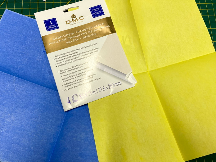 A white, gold, and blue package of transfer paper sits on top of two unfolded sheets of blue and yellow transfer paper.