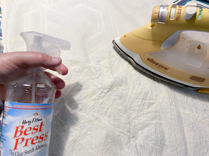 A hand is shown holding a bottle of Mary Ellen’s Best Press Starch Alternative spray over top of a wrinkled beige tea towel. A yellow Oliso Pro TG1600 Pro Plus Smart Iron is shown in the background.