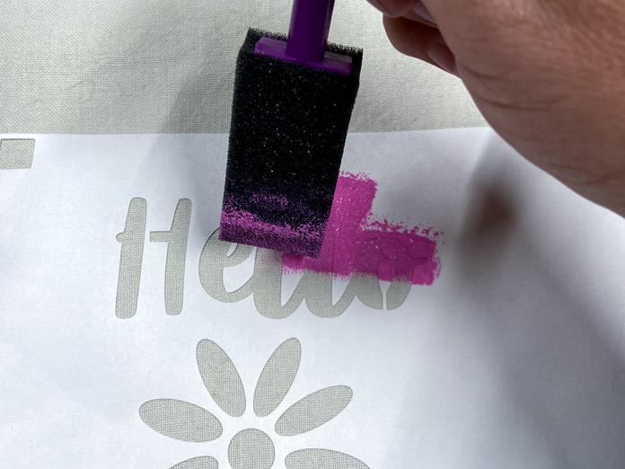 A hand holding a black foam craft brush dabs pink paint onto a stencil that has been ironed onto a beige tea towel; Mont Marte Signature Fabric Paints, Sew Easy Freezer Paper for Quilting and Applique