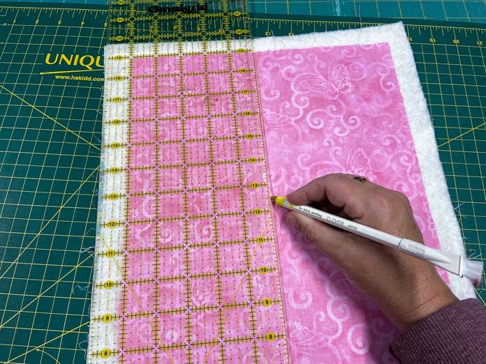 A hand is shown using a white marking pencil and rotary cutting ruler to draw quilting lines on the pink fabric. There is a green cutting mat in the background; Omnigrid ruler - 6" x 24", Clover 418 Chacopel Pencil Set – Fine Point