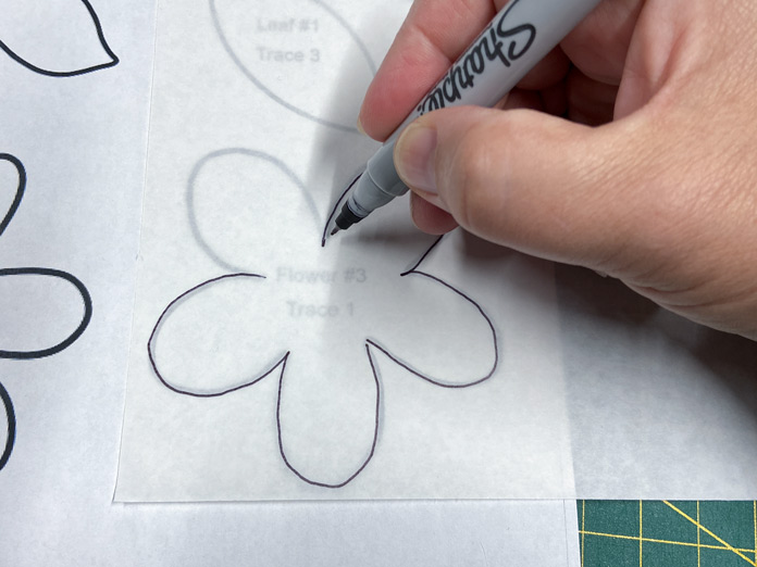 A hand holding a fine-tipped marker traces a flower design onto a sheet of HeatnBond EZ Print Feather Lite. A green cutting mat can be seen in the background.