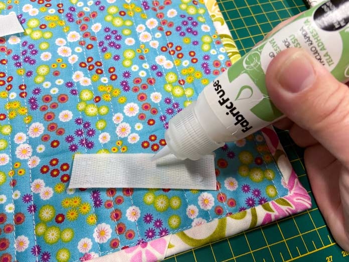 A hand is shown squeezing out small dots of adhesive from a small white and green bottle of Fabric Fuse by HeatnBond onto a white piece of hook and loop tape. The floral background of a small quilt is shown in the background; ESPRIT Hook and Loop Tape Sew-On - 19mm x 1m (3⁄4″ x 39″) – White