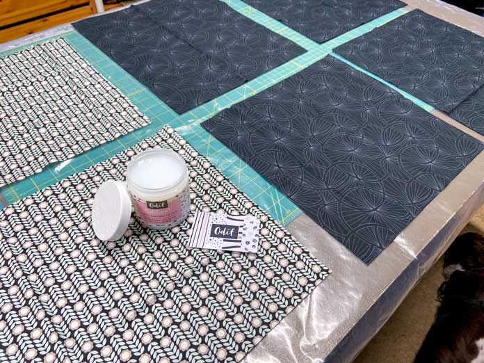 Four pieces of black fabric and two pieces of patterned fabric are shown lying on top of a plastic sheet on a cutting board. An open jar of Odif OdiCoat sits on top of one of the fabrics, with its lid leaning against it. The Odif applicator card sits beside the jar; Odif OdiCoat Gel Coating – 250ml, BEST PRESS Starch Alternative - 499mL (16.9 oz.) - Scent Free, BEST PRESS Spray and Misting Bottle - 295mL (10 fl. oz.) – Empty, Oliso Pro TG1600 Pro Plus Smart Iron, UNIQUE QUILTING Double Sided Applique Pressing Sheet - Brown - 46 x 46cm (18″ x 18″), Bosal In-R-Form Single Sided Fusible Foam Stabilizer - 45.7 x 147.3 cm (18″ x 58″), OLFA SCS-1 - 5″ Stainless Steel Serrated Edge Scissors, free quilting tutorials