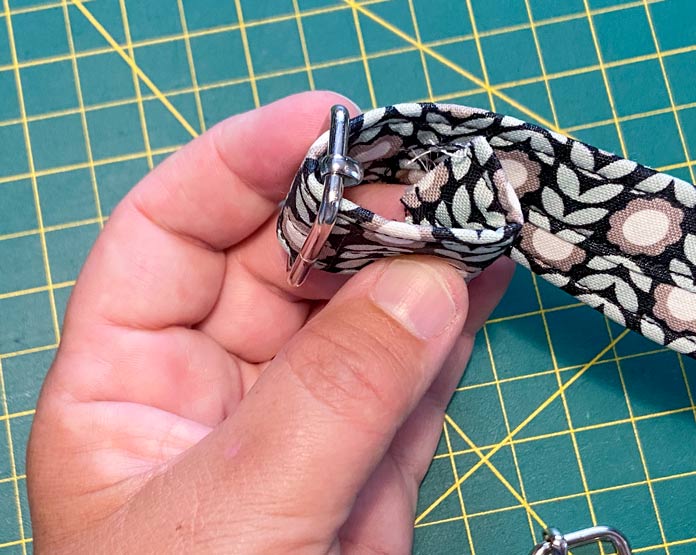 A hand is shown holding a silver buckle so that the folded under, strap end can be seen. A green cutting mat is in the background.