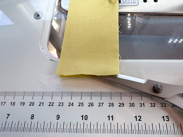 A yellow strip of fabric on the bed of a sewing machine is being cut at its fold with a pair of scissors. Hemline Gold Multi-Use Craft Bag (28.5cm x 36.5cm x 17.8cm), Hemline Gold Sewing Kit, Hemline Gold Quilters 4-in-1 Multi-Mat (30cm x 24cm), Hemline Gold Quilters Clips (Pack of 30), Hemlione Gold Retractable Tape Measure - 150cm/60, Oliso M3Pro Project Iron – Coral, OLFA 45mm RTY-2/DX/MAG Ergonomic Rotary Cutter – Magenta, OLFA RM-MG - 24″ x 36″ Double Sided Rotary Mat, UNIQUE Sewing Tools Holder, OLFA QR-6x12 - 6″ x 12″ Frosted Acrylic Ruler, OLFA QR-6x24 - 6″ x 24″ Frosted Acrylic Ruler, Fairfield Quilter's 80/20 Quilt Batting - 229 x 274cm (90″ x 108″)