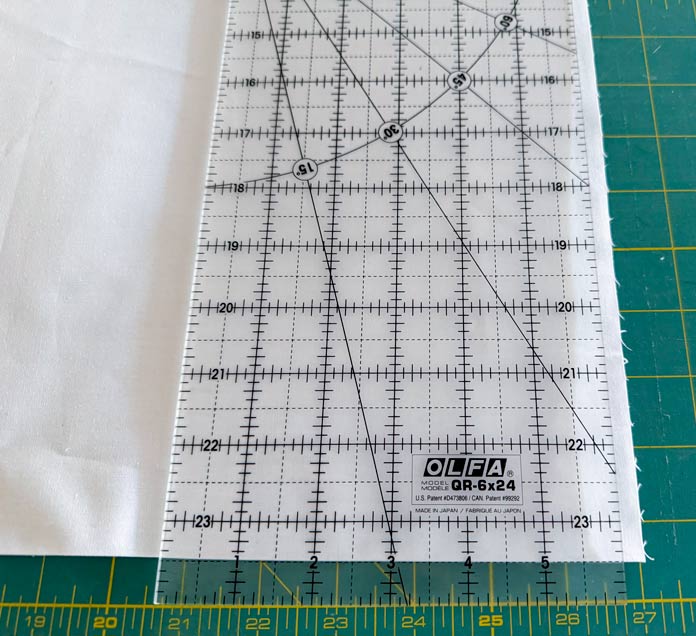 One line of an OLFA ruler is placed on the folded edge of white fabric on a green cutting mat. Hemline Gold Multi-Use Craft Bag (28.5cm x 36.5cm x 17.8cm), Hemline Gold Sewing Kit, Hemline Gold Quilters 4-in-1 Multi-Mat (30cm x 24cm), Hemline Gold Quilters Clips (Pack of 30), Hemline Gold Retractable Tape Measure - 150cm/60, oliso M3Pro Project Iron – Coral, OLFA 45mm RTY-2/DX/MAG Ergonomic Rotary Cutter – Magenta, OLFA RM-MG - 24″ x 36″ Double Sided Rotary Mat, UNIQUE Sewing Tools Holder, OLFA QR-6x12 - 6″ x 12″ Frosted Acrylic Ruler, OLFA QR-6x24 - 6″ x 24″ Frosted Acrylic Ruler, Fairfield Quilter's 80/20 Quilt Batting - 229 x 274cm (90″ x 108″).