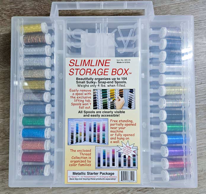 A clear plastic box with colored threads inside sits on a wood table. Husqvarna VIKING DESIGNER EPIC 3 sewing and embroidery machine, Husqvarna VIKING 6in/15.2cm Applique Scissor, Husqvarna VIKING Open Toe Presser Foot for IDF System, SULKY Slimline Metallic Dream Assortment