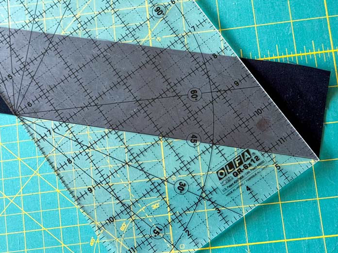 An OLFA quilting ruler marks a 45-degree angle line on the end of a black strip of fabric while on top of a green cutting mat. Hemline Gold Multi-Use Craft Bag (28.5cm x 36.5cm x 17.8cm), Hemline Gold Sewing Kit, Hemline Gold Quilters 4-in-1 Multi-Mat (30cm x 24cm), Hemline Gold Quilters Clips (Pack of 30), Hemline Gold Retractable Tape Measure - 150cm/60, oliso M3Pro Project Iron – Coral, OLFA 45mm RTY-2/DX/MAG Ergonomic Rotary Cutter – Magenta, OLFA RM-MG - 24″ x 36″ Double Sided Rotary Mat, UNIQUE Sewing Tools Holder, OLFA QR-6x12 - 6″ x 12″ Frosted Acrylic Ruler, OLFA QR-6x24 - 6″ x 24″ Frosted Acrylic Ruler, Fairfield Quilter's 80/20 Quilt Batting - 229 x 274cm (90″ x 108″) 