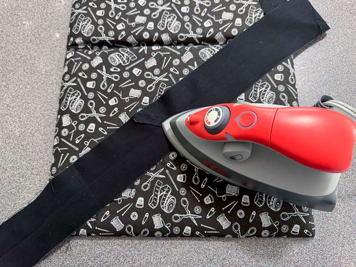 A black fabric strip is being pressed with a coral color oliso iron on a Hemline Gold Quilters 4-in-1 Multi-Mat with a black and white sewing print cover that doubles as an ironing surface. Hemline Gold Multi-Use Craft Bag (28.5cm x 36.5cm x 17.8cm), Hemline Gold Sewing Kit, Hemline Gold Quilters 4-in-1 Multi-Mat (30cm x 24cm), Hemline Gold Quilters Clips (Pack of 30), Hemline Gold Retractable Tape Measure - 150cm/60, oliso M3Pro Project Iron – Coral, OLFA 45mm RTY-2/DX/MAG Ergonomic Rotary Cutter – Magenta, OLFA RM-MG - 24″ x 36″ Double Sided Rotary Mat, UNIQUE Sewing Tools Holder, OLFA QR-6x12 - 6″ x 12″ Frosted Acrylic Ruler, OLFA QR-6x24 - 6″ x 24″ Frosted Acrylic Ruler, Fairfield Quilter's 80/20 Quilt Batting - 229 x 274cm (90″ x 108″) 