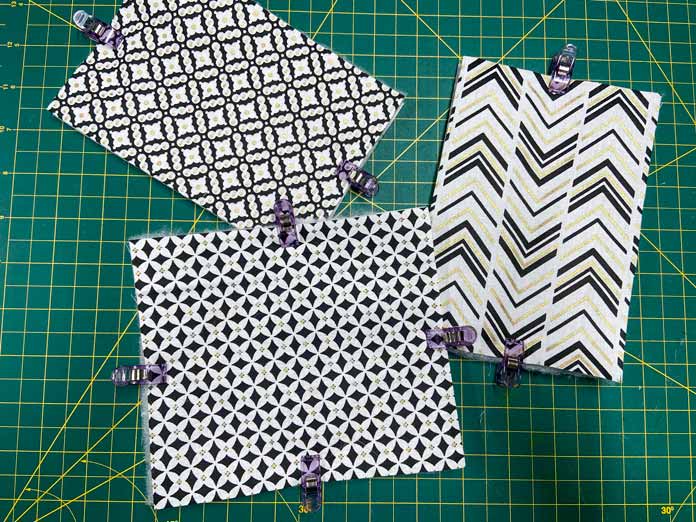 Three rectangular quilt sandwiches made with white and black fabric sit on a green cutting mat. Purple clips keep the layers together. Oliso M2Pro Mini Project Iron, FAIRFIELD Low-Loft Quilt Batting - Crib - 114 x 152cm (45″ x 60″), UNIQUE QUILTING Clever Clips, OLFA CHN-1 - Chenille / Textile Cutter, UNIQUE CREATIV Trim Pack - Black & White, FABRIC CREATIONS Burlap Fabric - Oyster - 45 x 61cm (18″ x 24″), FABRIC CREATIONS Faux Fur Fabric - Zebra - 45 x 76cm (18″ x 30″), UNIQUE SEWING Double Adjust Parachute Buckle - Plastic - 25mm (1″) White, CRAFTING ESSENTIALS Bottle of Buttons - Pink Tones - 75g (2.6oz), UNIQUE SEWING Invisible Magnetic Snaps - Sew-On - 12mm (1⁄2″) - 2 sets, UNIQUE QUILTING Stitch-N-Steam - 157 x 45cm (62″ x 18″), FABRIC CREATIONS Fabric Bundle (5pcs) - Metallic Gold - 45 x 53cm (18in x 21in), Oliso Pro TG1600 Pro Plus Smart Iron, UNIQUE QUILTING Clever Clips Large - 12pcs, sewing tutorial