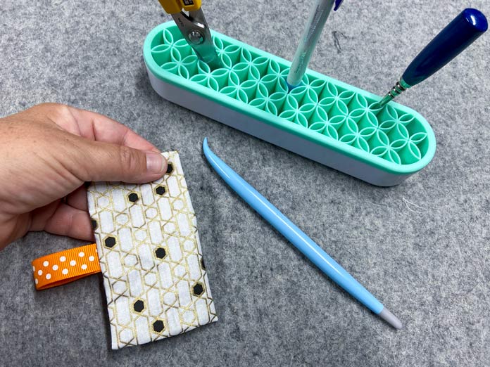 A hand holds a flap of fabric that has an orange ribbon attached to it. A grey wool pressing mat is in the background, and a blue stiletto and other tools sit nearby. OLFA CHN-1 - Chenille / Textile Cutter, UNIQUE QUILTING Stitch-N-Steam - 157 x 45cm (62″ x 18″), FABRIC CREATIONS Fabric Bundle (5pcs) - Metallic Gold - 45 x 53cm (18in x 21in), UNIQUE SEWING Self-Gripping Fastener Strip - 25cm (10″) - White, UNIQUE Webbing 25mm x 10m (1″ x 11yd) - White, an ELAN Trench Buckle - 45mm (13⁄4″) - Brown, UNIQUE CREATIV Trim Pack - Black & White, UNIQUE QUILTING Clever Clips Small - 12 pcs, CLOVER 7807 - Hold it Precision Stiletto, UNIQUE SEWING Sewing Tools Holder, UNIQUE QUILTING Wool Pressing Mat - 18″ x 24″ - Grey, OLISO M2Pro Mini Project Iron with Solemate, UNIQUE QUILTING Clever Clips Large - 12pcs, UNIQUE SEWING Invisible Magnetic Snaps - Sew-On - 12mm (1⁄2″) - 2 sets, COSTUMAKERS Activewear One Way Separating Zipper 23cm (9″) - White - 1760, UNIQUE SEWING Double Adjust Parachute Buckle - Plastic - 25mm (1″) - White, free tutorial