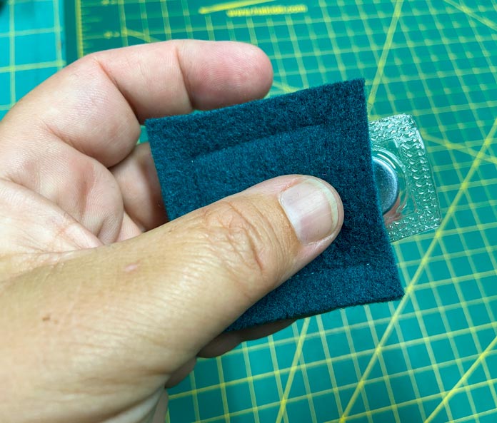 A hand is holding a square of dark green wool felt with a magnet poking out the side. A green cutting mat can be seen in the background. UNIQUE SEWING Self-Gripping Fastener Strip - 25cm (10″) - White, UNIQUE Webbing 25mm x 10m (1″ x 11yd) - White, ELAN Trench Buckle - 45mm (13⁄4″) - Brown, UNIQUE CREATIV Trim Pack - Black & White, UNIQUE SEWING Invisible Magnetic Snaps - Sew-On - 12mm (1⁄2″) - 2 sets,  SOFTKUT Pinking Shears (Scissors) - 81⁄2″ (21.6cm), COSTUMAKERS General Purpose Closed End Zipper 20cm (8″) - White - 1700, UNIQUE CREATIV Trim Pack - Red & Green, OLFA RTY-2/DX - Deluxe Ergonomic Handle Rotary Cutter 45mm, UNIQUE QUILTING Clever Clips Small - 12 pcs, UNIQUE SEWING Double Adjust Parachute Buckle - Plastic - 25mm (1″) - White, CRAFTING ESSENTIALS Bottle of Buttons - Pink Tones - 75g (2.6oz), UNIQUE Braided Elastic 3mm x 3.7m - White, free tutorial