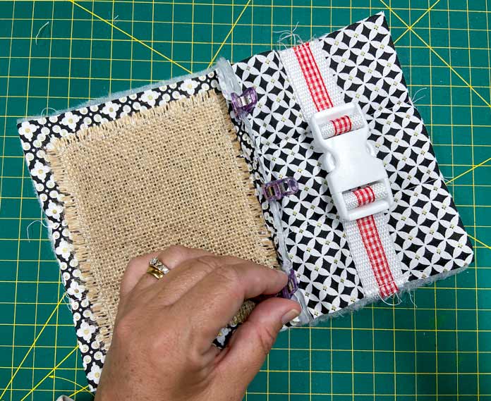 A hand is holding a small purple clip attached to the seam allowance between 2 quilt blocks, 1 with a burlap top and the other with a parachute buckle. A green cutting mat is in the background. UNIQUE SEWING Invisible Magnetic Snaps - Sew-On - 12mm (1⁄2″) - 2 sets, COSTUMAKERS General Purpose Closed End Zipper 20cm (8″) - White - 1700, UNIQUE SEWING Double Adjust Parachute Buckle - Plastic - 25mm (1″) – White, UNIQUE QUILTING Clever Clips Small - 12 pcs, CLOVER 7812 Roll & Press, UNIQUE QUILTING Wool Pressing Mat - 18″ x 24″ - Grey, OLISO M2Pro Mini Project Iron with Solemate, CLOVER 240 - Fork Pins - .56mm, CLOVER 464/18 - Bias Tape Maker - 18mm (3⁄4″), Odif 505 Temporary Quilt Basting Adhesive Fabric Spray - 156g, FAIRFIELD Low-Loft® Quilt Batting - Crib - 114 x 152cm (45″ x 60″), free tutorial