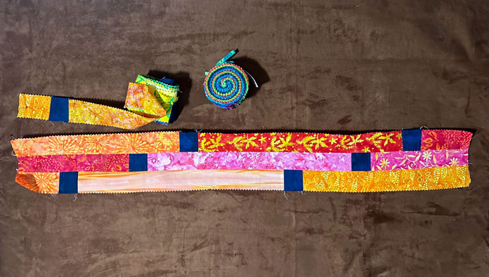 3 bright colored strips of batik serged together with a 4-thread overlock seam, in 20’ sections with a dark colored square separating each, shown with a pieced roll ready to add to the previously pieced rows. Also shown is the original jelly roll; PFAFF admire air 7000 serger 