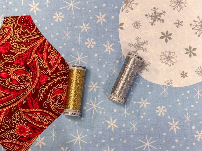 A spool of gold metallic thread is next to a red and gold fabric and a spool of silver metallic thread is next to a grey and white fabric with a light blue snowflake fabric in the background; Gütermann Dekor Metallic Thread 200m – Gold, Gütermann Dekor Metallic Thread 200m - Col. 41, Gütermann Dekor Metallic Thread 200m – Red, Gütermann Dekor Metallic Thread 200m - Christmas Green, Gütermann Dekor Metallic Thread 200m - Col. 1005, Gütermann Denim Thread 100m – Black, UNIQUE Baby Rick Rack 3mm x 3.7m – Scarlet, UNIQUE Medium Rick Rack 6mm x 2.3m - Emerald Green, UNIQUE Jumbo Rick Rack 10mm x 2.3m - Metallic Gold, SCHMETZ #1743 Metallic Needles Carded - 80/12 - 5 count, SCHMETZ #1752 Metallic Needles Carded - 90/14 - 5 count, LDH 8½“ Craft Scissors – Purple, HeatnBond Lite Iron-On Adhesive - 43cm x 30m (17″ x 32 ¾yds) bolt, HeatnBond Stitch n Sew Tear-Away Stabilizer - 50.8cm x 22.8m (20″ x 25yds), oliso M3Pro Project Iron – Coral, OLFA 45mm RTY-2/DX/MAG Ergonomic Rotary Cutter – Magenta, UNIQUE Quilting Wool Pressing Mat - 18″ x 24″ - Grey