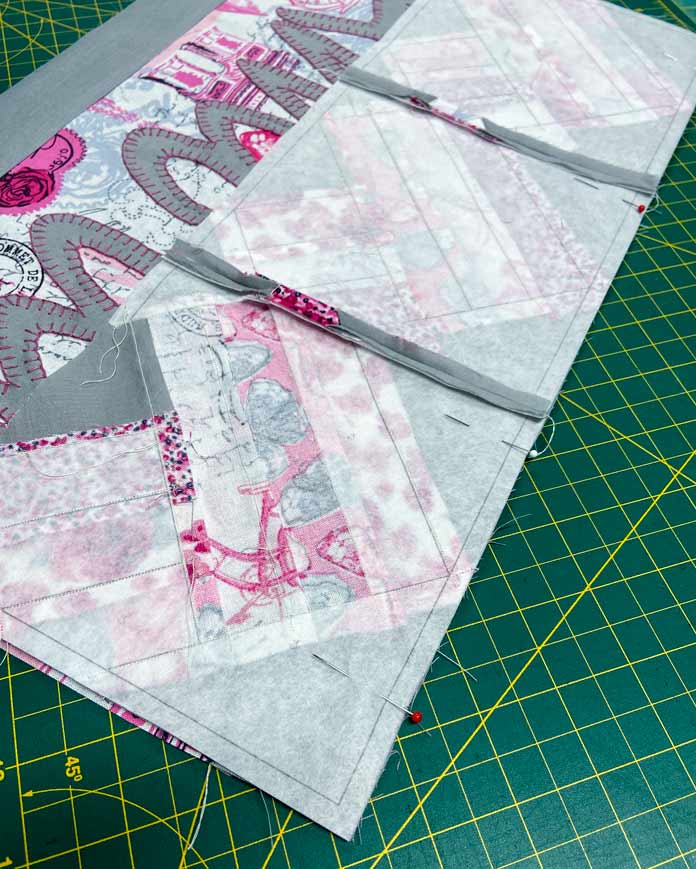 A pink, gray and white fabric rectangle with the word Amour appliqued on top sits on a green cutting mat. A row of paper pieced blocks has been pinned to the side using red and white pins. 