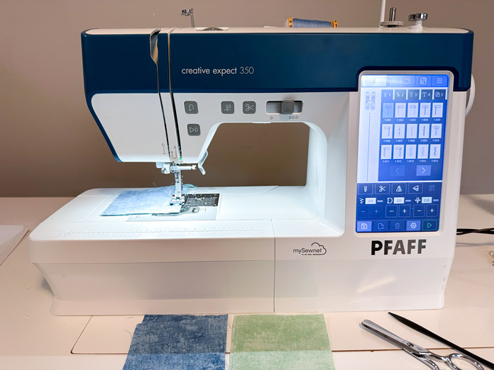 A small white sewing machine setup with blue cotton thread and a ¼” piecing foot showing blue and green squares being pieced together; PFAFF creative expect 350, sewing machine reviews, mySewnet Quilting Software, mini quilt finishing, beginner quilting tips, quilt binding guide, machine quilting techniques, organic quilting, embroidery quilt labels, diy quilt projects, free motion quilting, free quilting project, free quilting patterns, baby quilt, free quilting tutorials