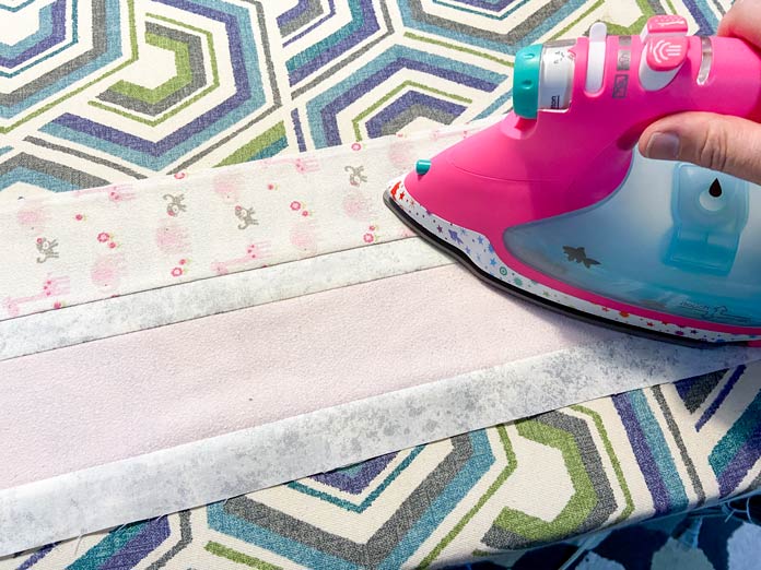 A hand uses a bright pink iron to press the seams of a rectangle of fabrics that have been sewn together. They sit on a multicolored ironing board cover.