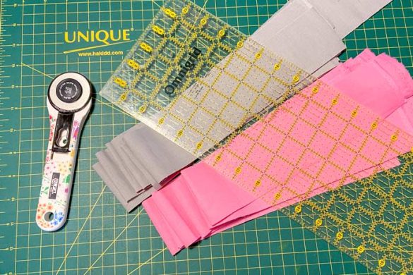 A white rotary cutter with flower decorations on the handle sits beside a pile of gray fabric strips and a pile of pink fabric strips. A rotary cutting ruler sits on top of the strips and a green cutting mat is in the background.