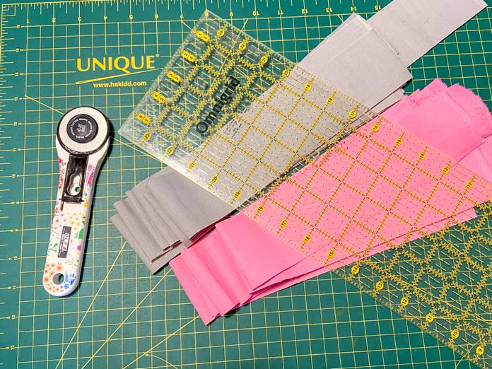 A white rotary cutter with flower decorations on the handle sits beside a pile of gray fabric strips and a pile of pink fabric strips. A rotary cutting ruler sits on top of the strips and a green cutting mat is in the background.