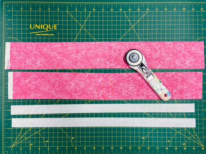 Two narrow strips of white interfacing and two wider strips of pink fabric sit on a green cutting mat. A white rotary cutter with a flower design on the handle sits on top of the strips.
