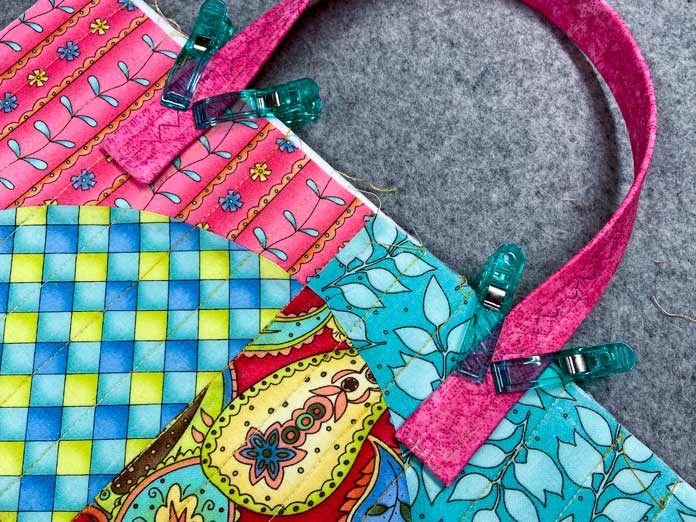 A pink handle is clipped to a multicolored quilted panel using large green clips. A grey wool pressing mat sits in the background.