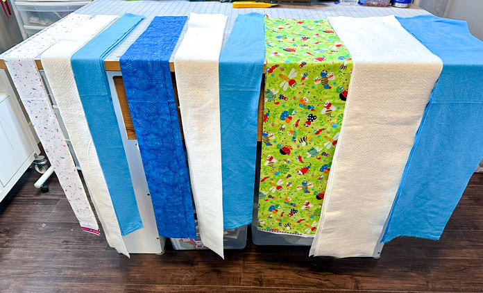 Cutting board with piles of 3” white print fabric, batting and backing (blue flannel), 4.5” blue batik, batting, and backing, and 9” green bug print fabric, batting and backing. All laid out in order of use.