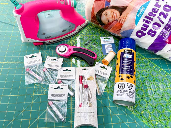 Tools for the week, an Oliso Pro Smart Iron, OMNIGRIP ruler, OLFA 45mm Ergonomic Rotary Cutter, SCHMETZ Universal Needles 80/12, Gütermann Cotton 50wt Thread Ivory, BOHIN Mechanical Chalk Pencil 0.9mm, BOHIN Pencil Refills (Pink, Grey, Green, Yellow and White, Odif 505 Temporary Quilt Basting Adhesive, FAIRFIELD Quilter's 80/20 Quilt Batting, all on an OLFA 24″ x 36″ Double Sided Rotary Mat