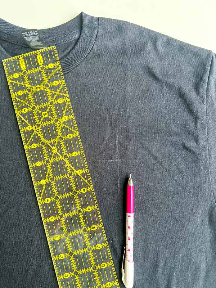 T-shirt with center cross marked using a white mechanical fabric pencil, positioned under a yellow quilting ruler for precise alignment. This is the first step in preparing the garment for embroidery on the PFAFF creative icon 2 sewing and embroidery machine