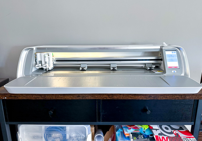 A 24” wide white and grey digital craft cutting machine, with the front door open, lights on, showing a small computer screen on the right and a computer-controlled rotary blade installed on the left.