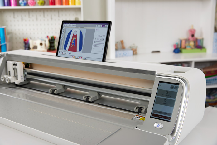 A 24” wide white and gray digital craft cutting machine, with the front door open, lights on, showing a small computer screen on the right and a computer-controlled rotary blade installed on the left.