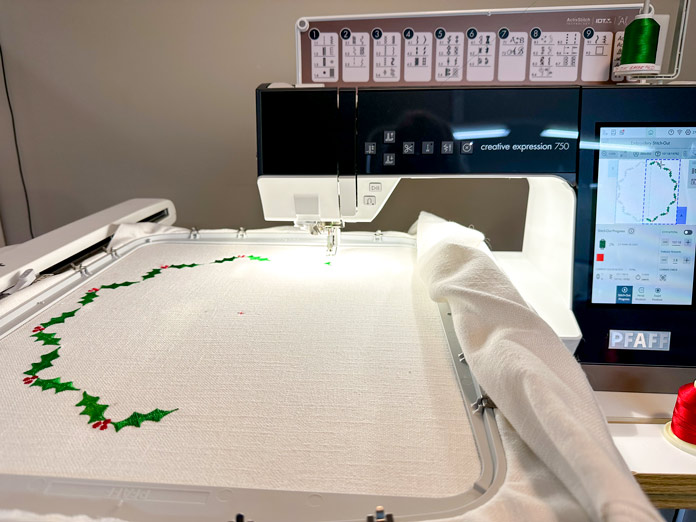 A picture of a deep charcoal and white sewing /embroidery machine with a large computer screen, turned on, waiting to be used. A white embroidery arm attached, and a large 360X350 hoop attached with a cream piece of fabric inserted