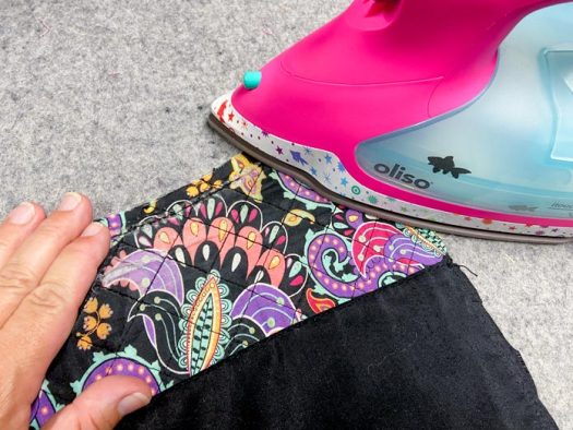 A hand is shown holding the corner of a multicolored purse while a pink iron presses the edge on a grey wool pressing mat.