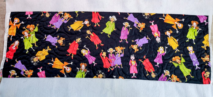 Black fabric with colorful choir ladies on a larger piece of white fleece; Husqvarna Viking Designer Ruby 90 sewing and embroidery machine, Inspira Fusible Fleece, Omnigrip 20½" square ruler, Singer 20 Steam Garment Press