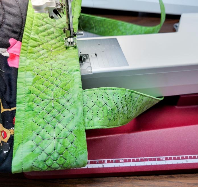 Green fabric embellished with machine embroidery under the free arm of a computerized sewing machine; Husqvarna Viking Designer Ruby 90 sewing and embroidery machine, Husqvarna Viking Extension Table with Adjustable Guide, Inspira Top Stitch Needles, Husqvarna Viking Multi-Function Foot Control, Inspira Fusible Fleece Stabilizer