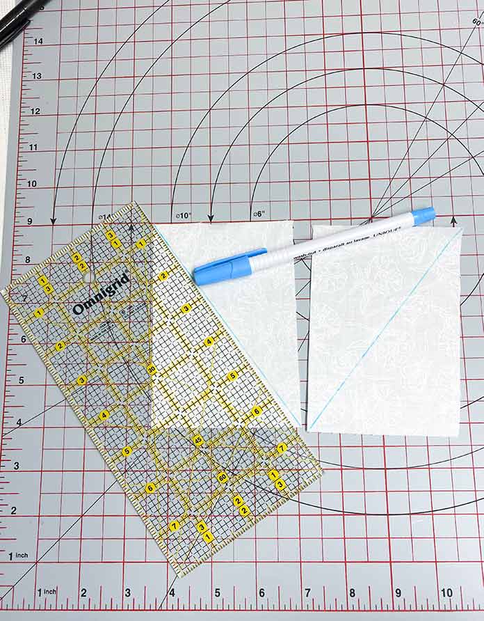 A quilting ruler, a marking pen, and two white fabric rectangles with blue diagonal drawn lines, sitting on a grey cutting mat. SCHMETZ Quilting needles, UNIQUE Sewing Fast Fade Fabric Markers, Omnigrid Rulers, OLFA Rotary Cutter, Komfort Kut Rotating Cutting mat, Fabric Creations Cotton Fabric.