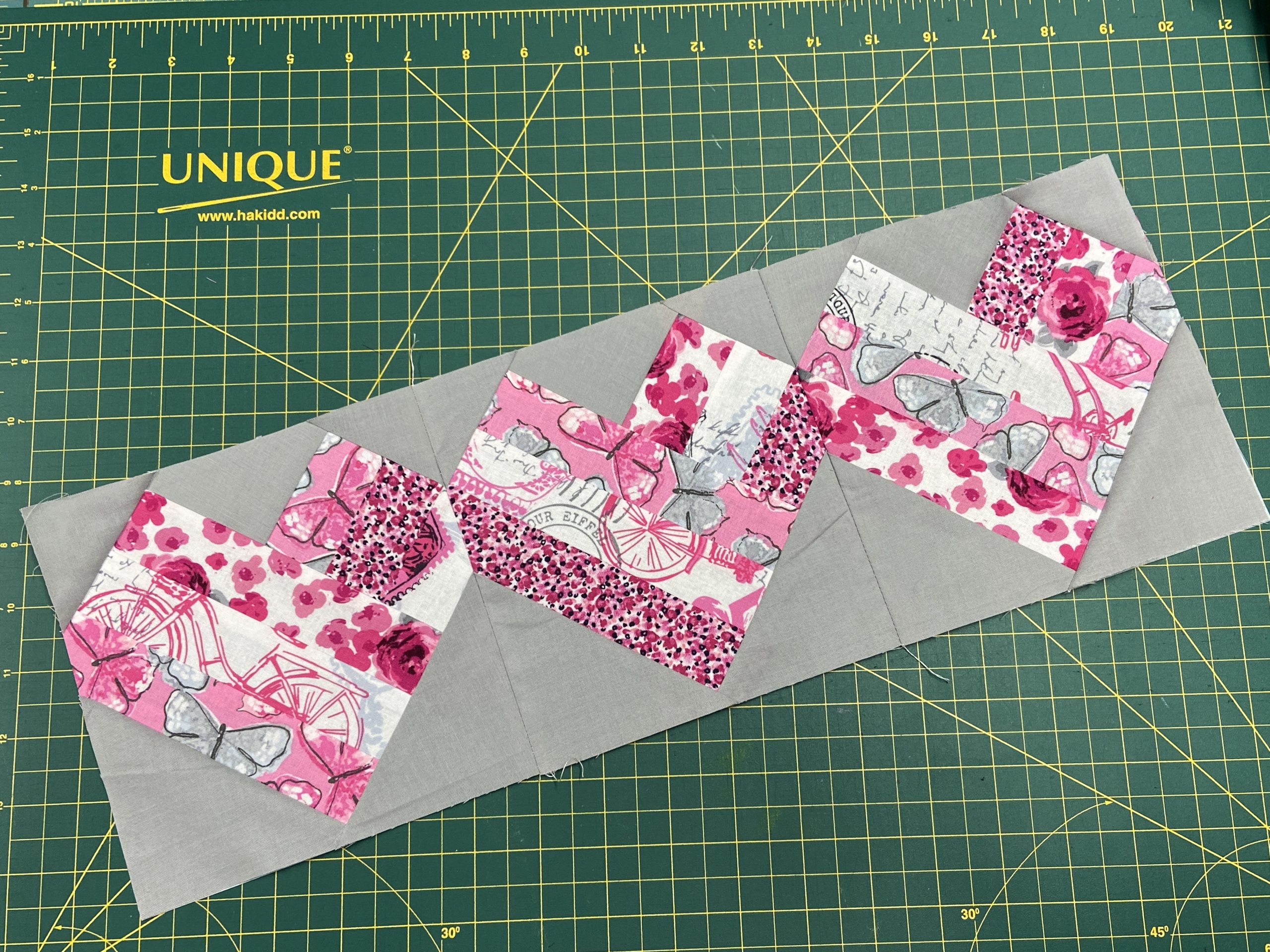 A row of pink and gray heart quilt blocks sits on top of a green cutting board.